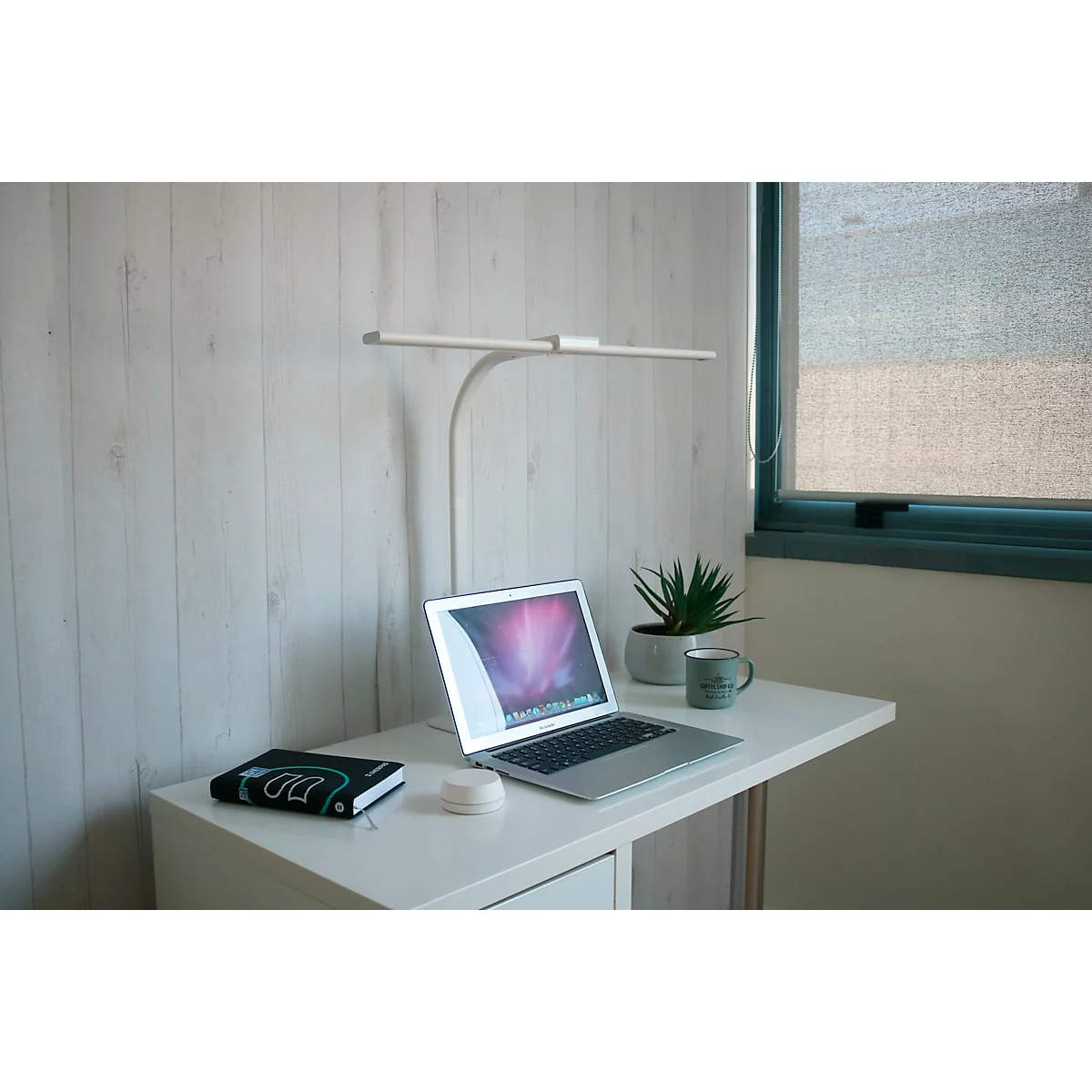 Schreibtisch mit Laptop, Lampe, Pflanze und Notizbuch. Am Fenster hängt ein Rollo.