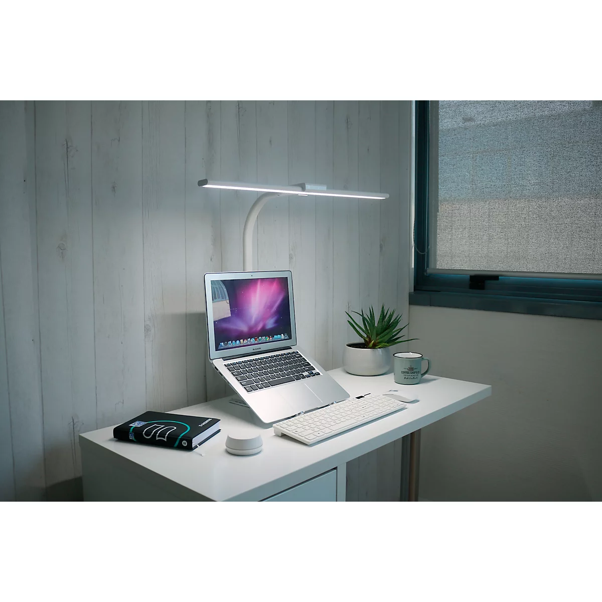 Schreibtisch mit Laptop, Tastatur, Lampe und Topfpflanze. Die Lampe scheint hell. Das Fenster lässt natürliches Licht herein.