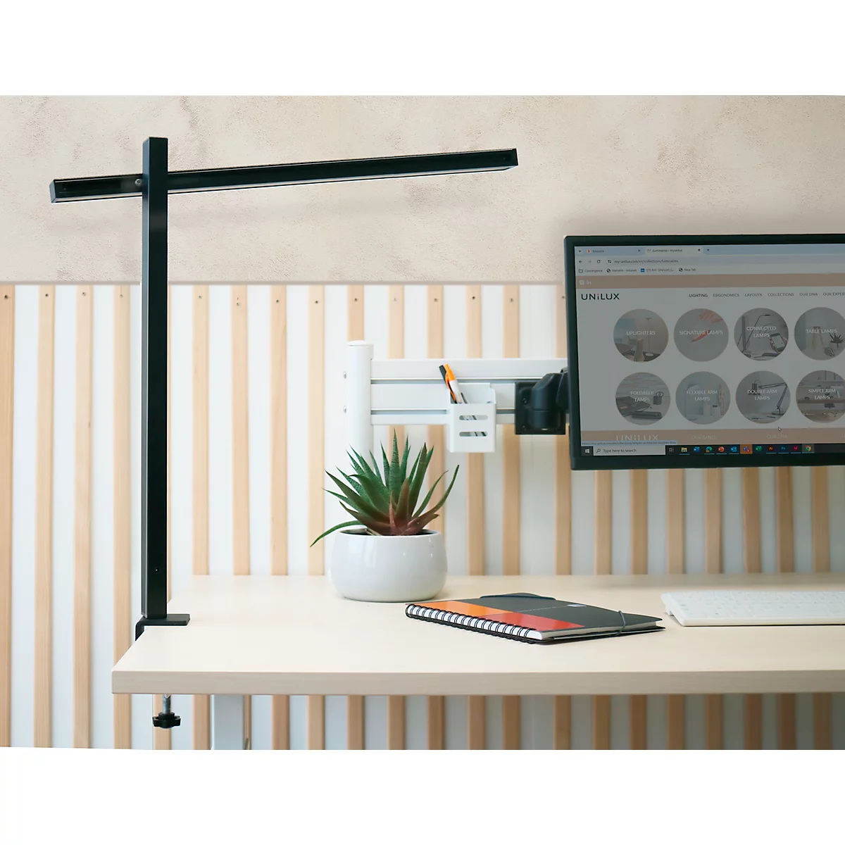 Schwarze Schreibtischlampe, Monitor, Pflanze, Notizblock auf einem hellen Schreibtisch. Im Hintergrund Holzvertäfelung.
