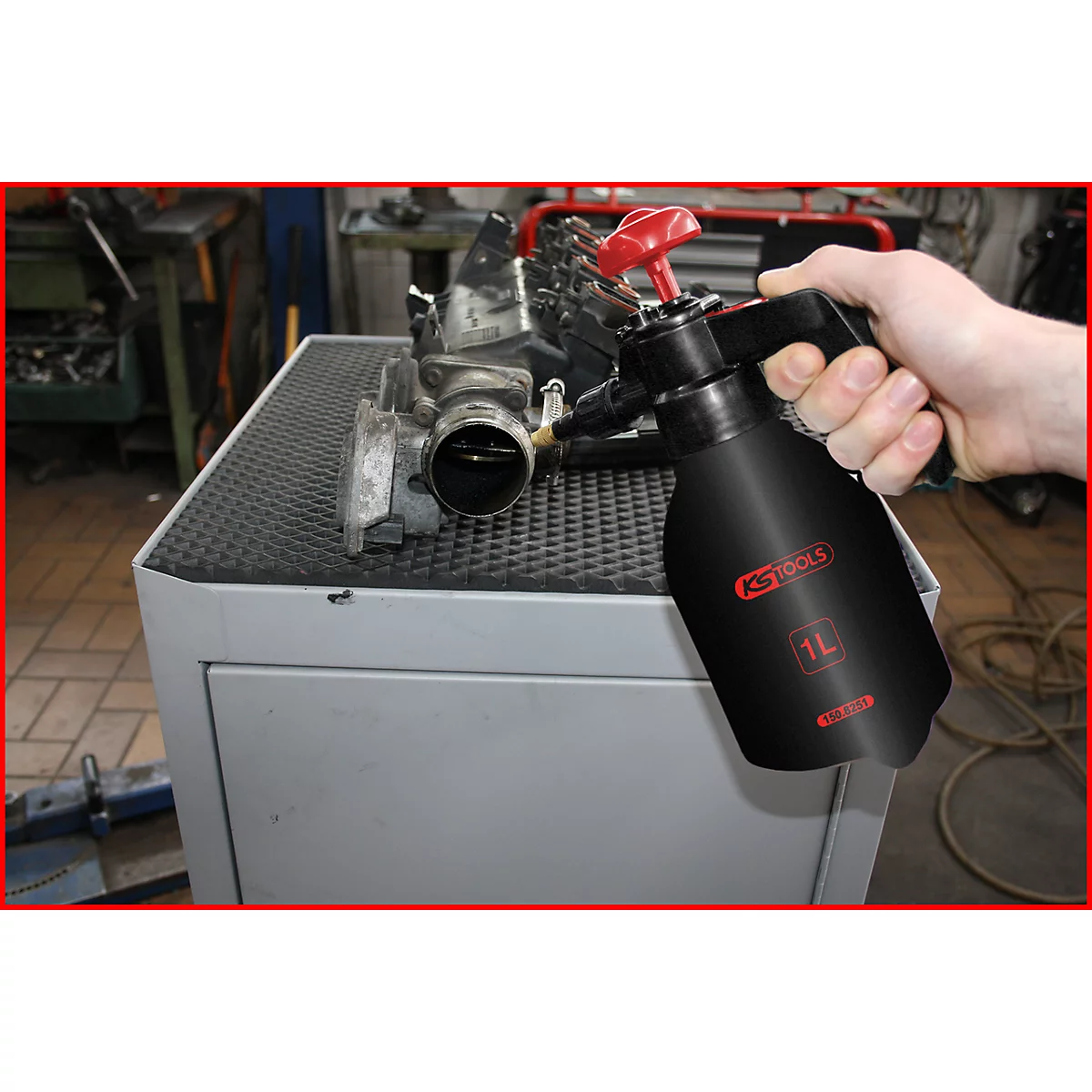 Eine schwarze Sprühflasche mit rotem Aufsatz, gehalten von einer Hand, vor einem Motorteil. Aufschrift: KS TOOLS, 1L.