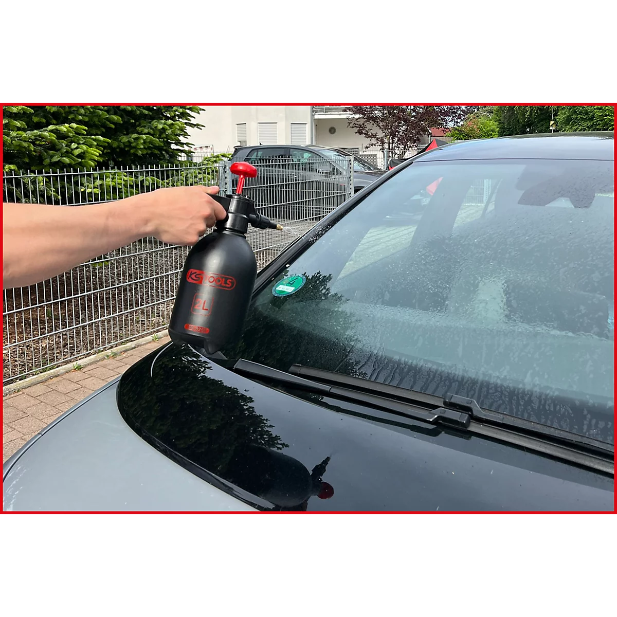 Person reinigt eine Autoscheibe mit einer schwarzen Sprühflasche. Die Scheibe ist feucht und es ist ein Auto im Hintergrund zu sehen.