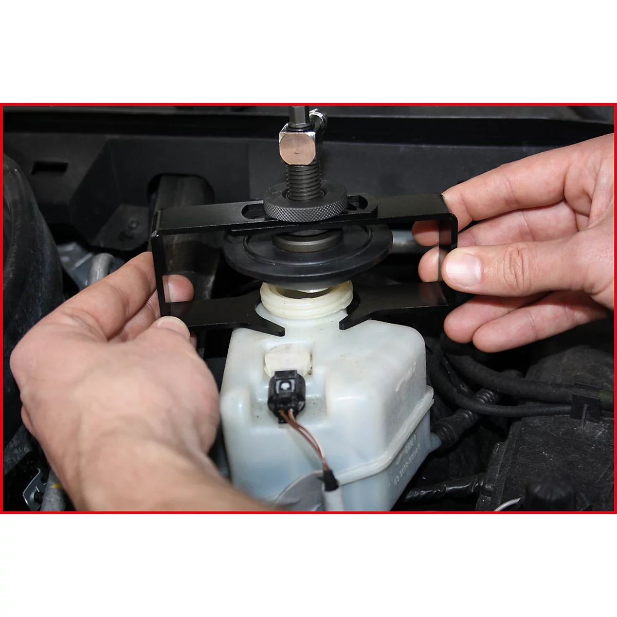 Hände halten ein schwarzes Werkzeug über einem Behälter im Motorraum eines Autos. Das Werkzeug hat eine Schraube und eine Halterung.