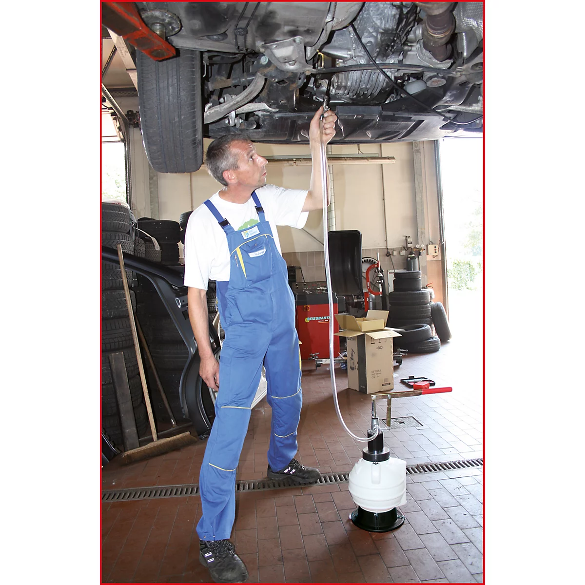 Ein Mechaniker in blauer Arbeitskleidung arbeitet unter einem Auto. Er hält einen Schlauch zu einer weißen Flüssigkeitsabsauganlage.