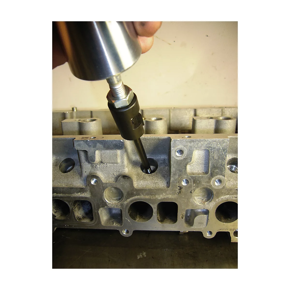 Eine Hand hält ein Werkzeug, das in ein Loch in einem grauen Metallteil gesteckt wird. Der Hintergrund ist unscharf.