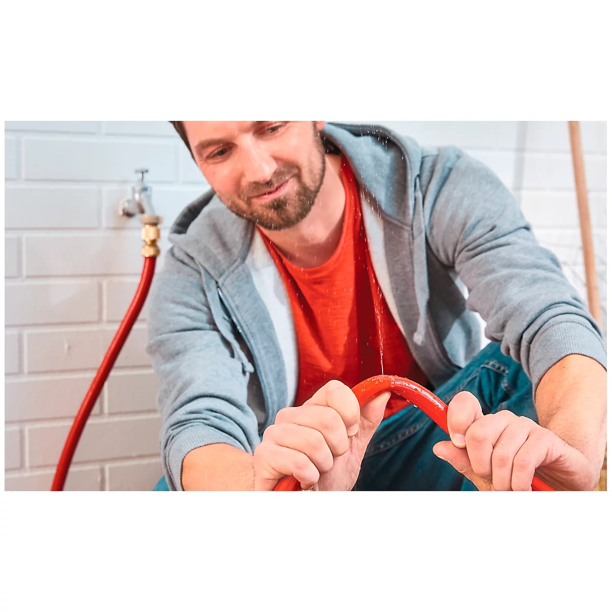 A man repairs a red water hose. He looks concentrated, face with a beard. In the background a faucet and a white wall.