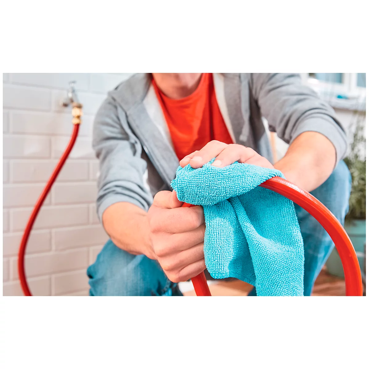 Man cleaning a red hose with a blue cloth, a faucet and tiles in the background.