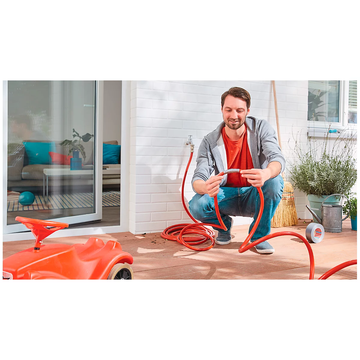 Man crouching, connecting red garden hose. In the background: house, toy car.