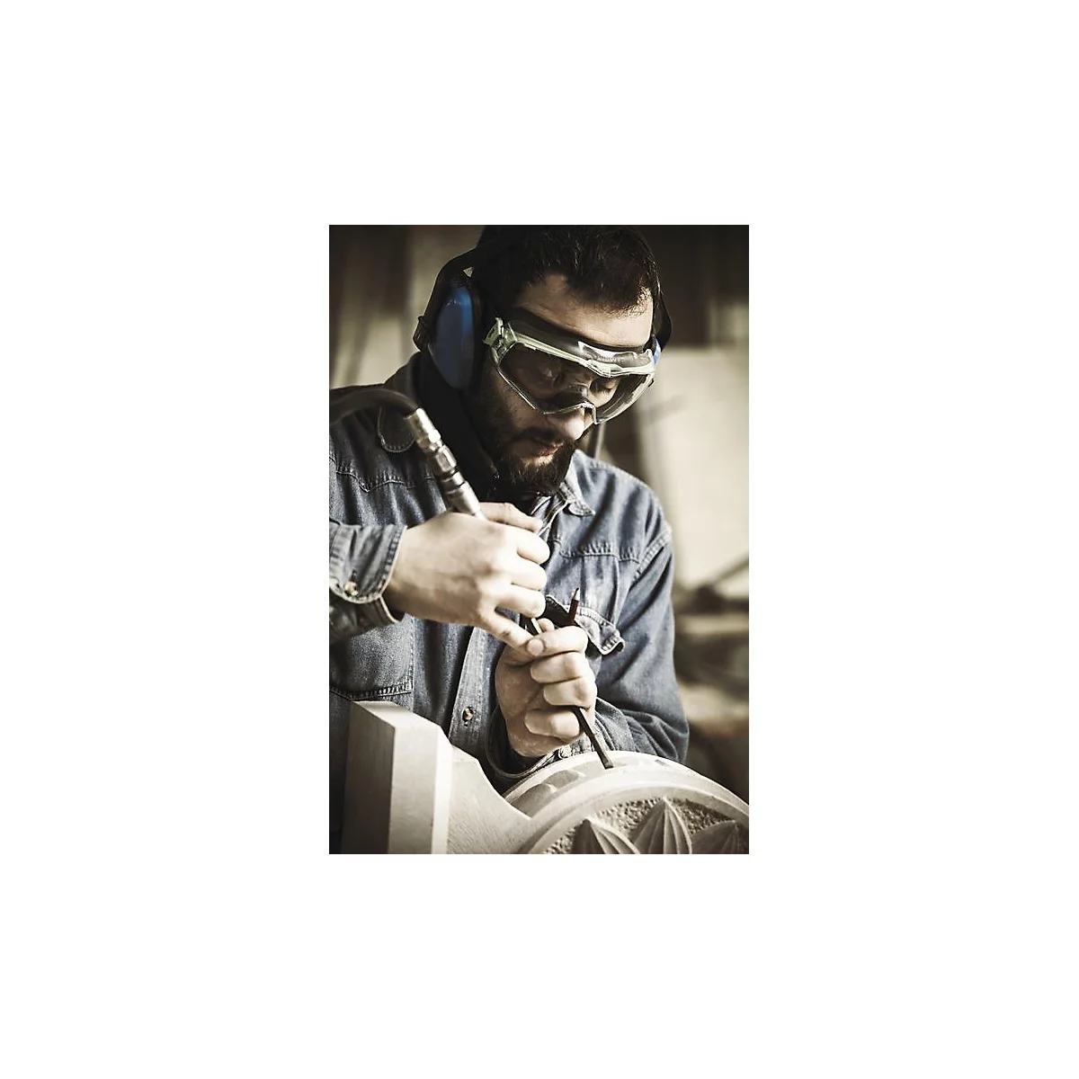 Un homme sculpte la pierre avec des lunettes de sécurité et une protection auditive. Il travaille concentré sur une œuvre d'art ornée.