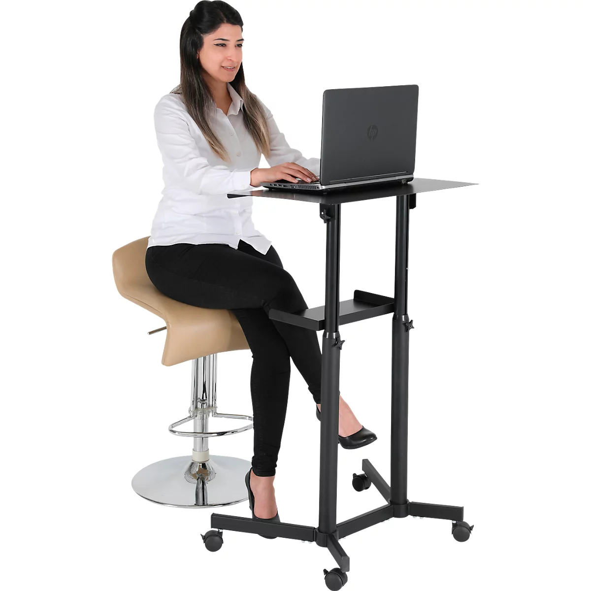 A woman sits at a height-adjustable standing desk with a laptop. She wears a white shirt and black trousers.