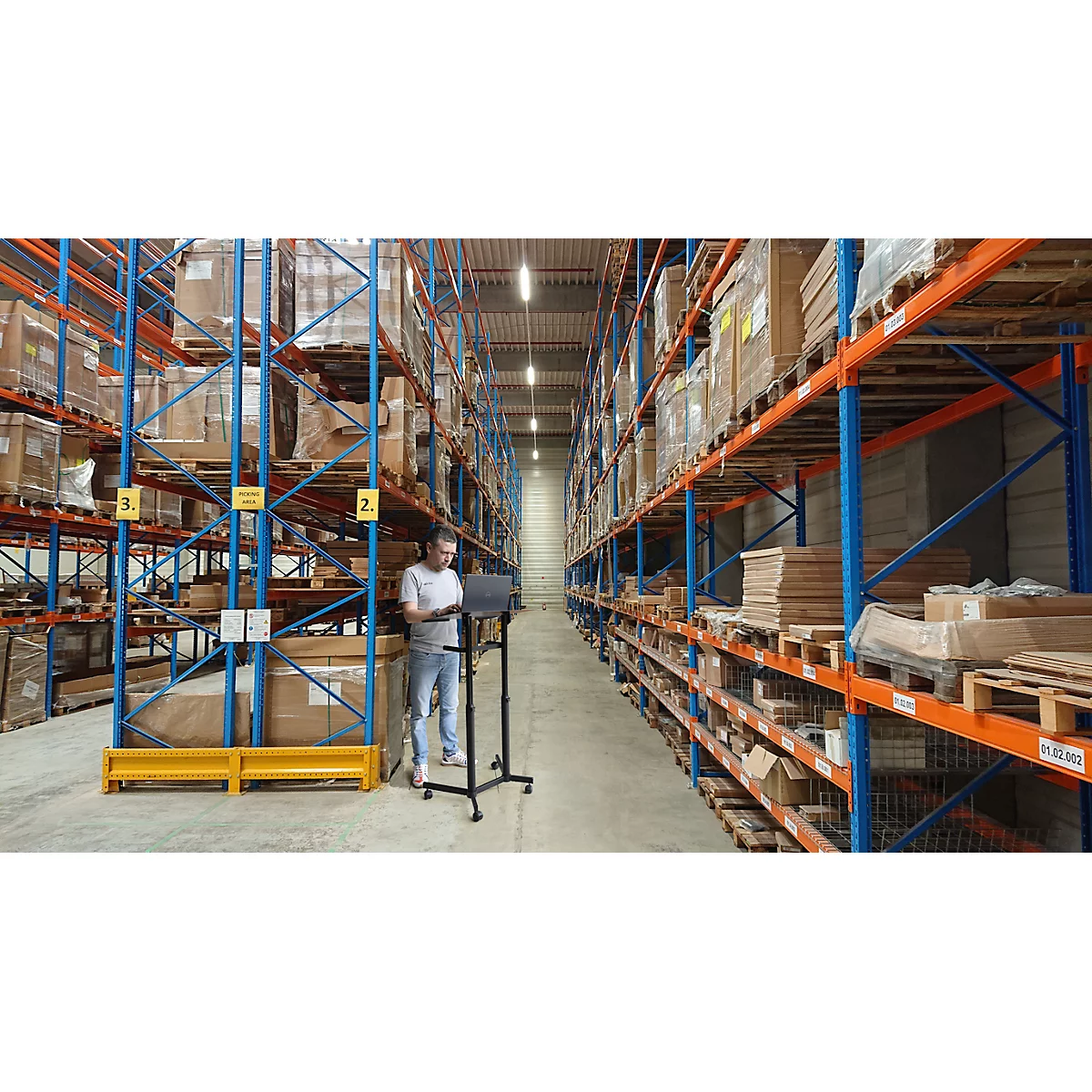 Man with a laptop in a warehouse between racks of boxes. Blue and orange metal shelving.
