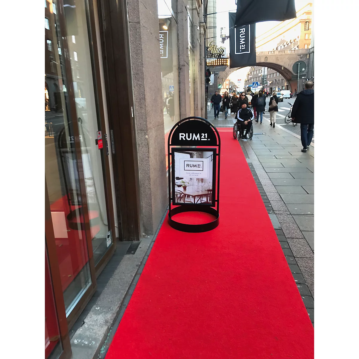 Tapis rouge devant un magasin. Un panneau publicitaire avec "RUM21" et des piétons en arrière-plan.