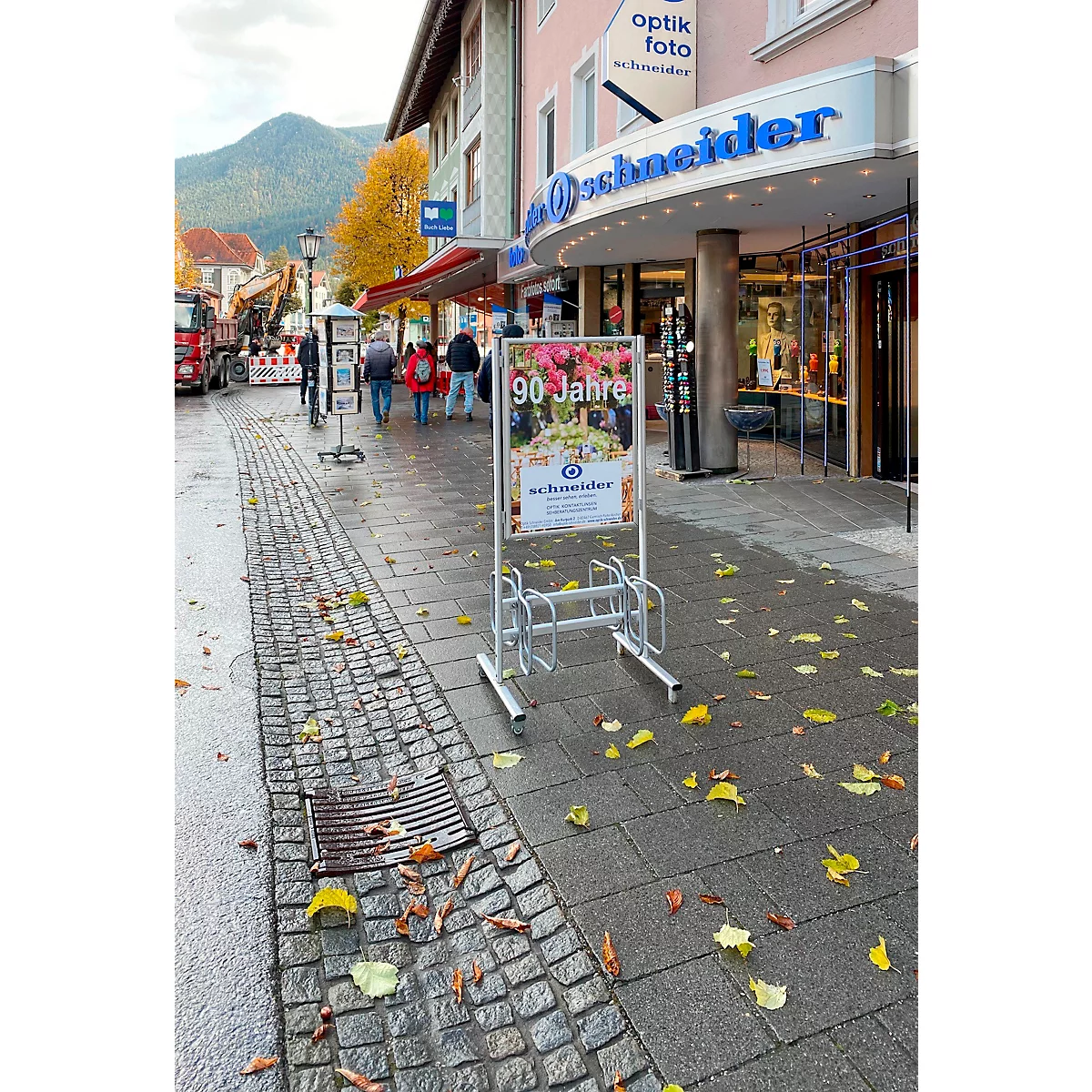 Support à vélos devant un magasin avec l'inscription "Optik Foto Schneider" et une enseigne publicitaire pour un 90e anniversaire.