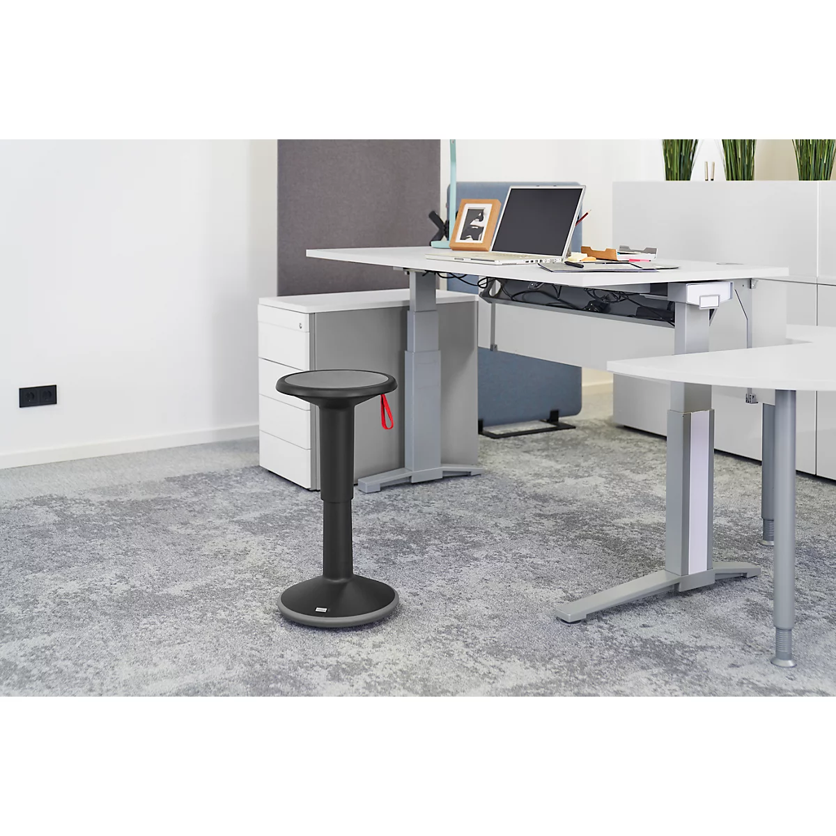 Office stool in front of height-adjustable desk. Desk with laptop, plants. Gray carpet, light walls.