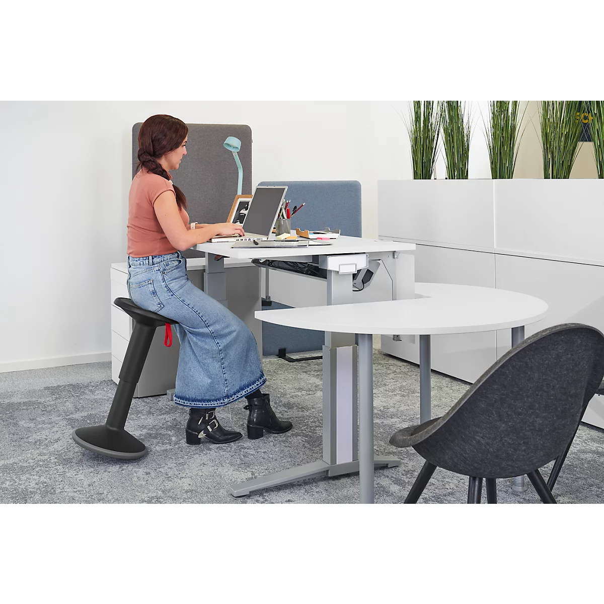 A woman is sitting at a desk working on a laptop. She is wearing jeans and boots.