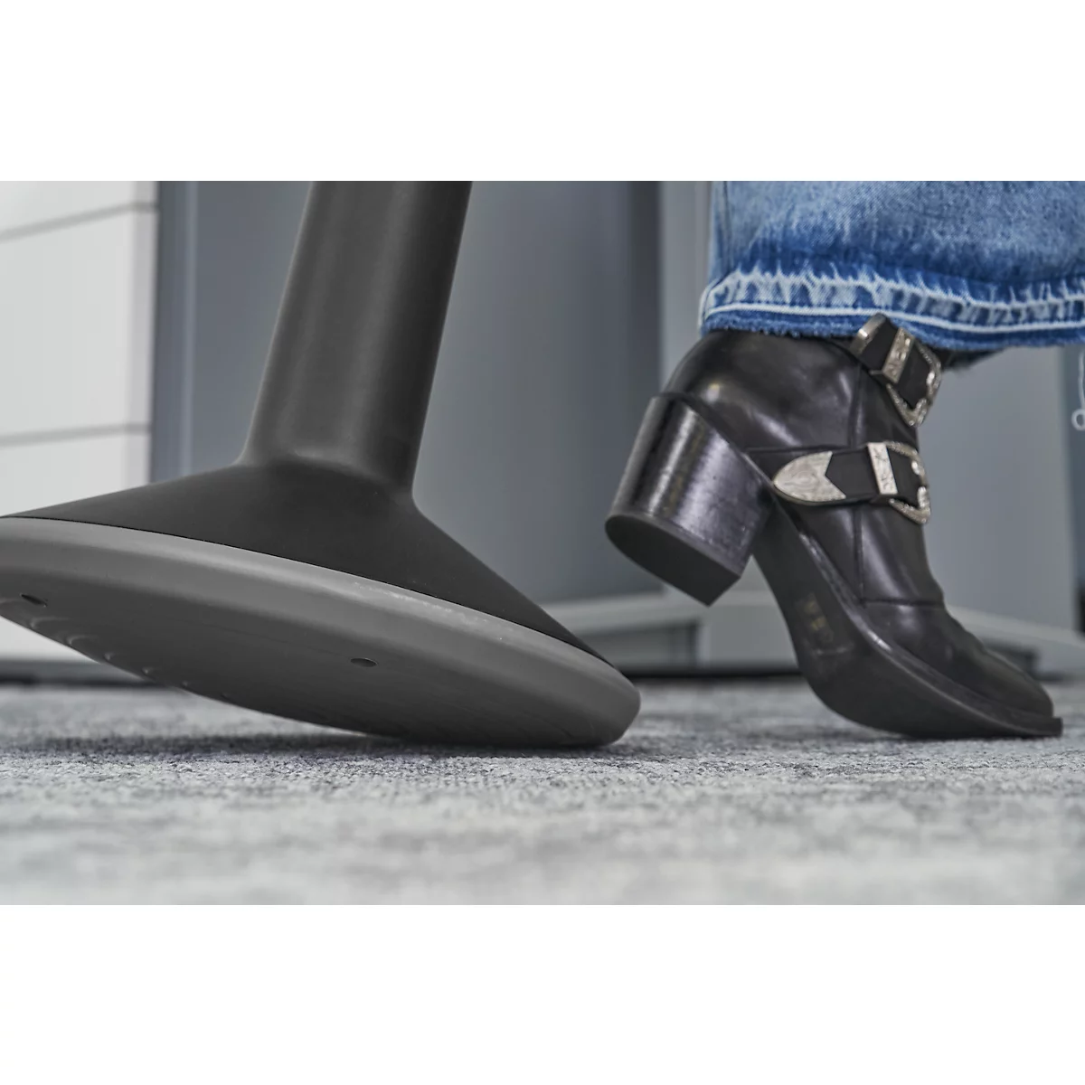 Black leather shoe with silver buckle and a chair leg. The shoe is visible on the foot. In the background a grey carpet.