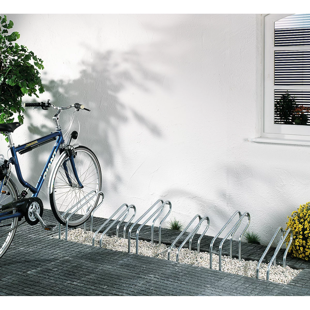 Blue bicycle in a bike rack. The rack is mounted on a paved path in front of a white wall with a window.