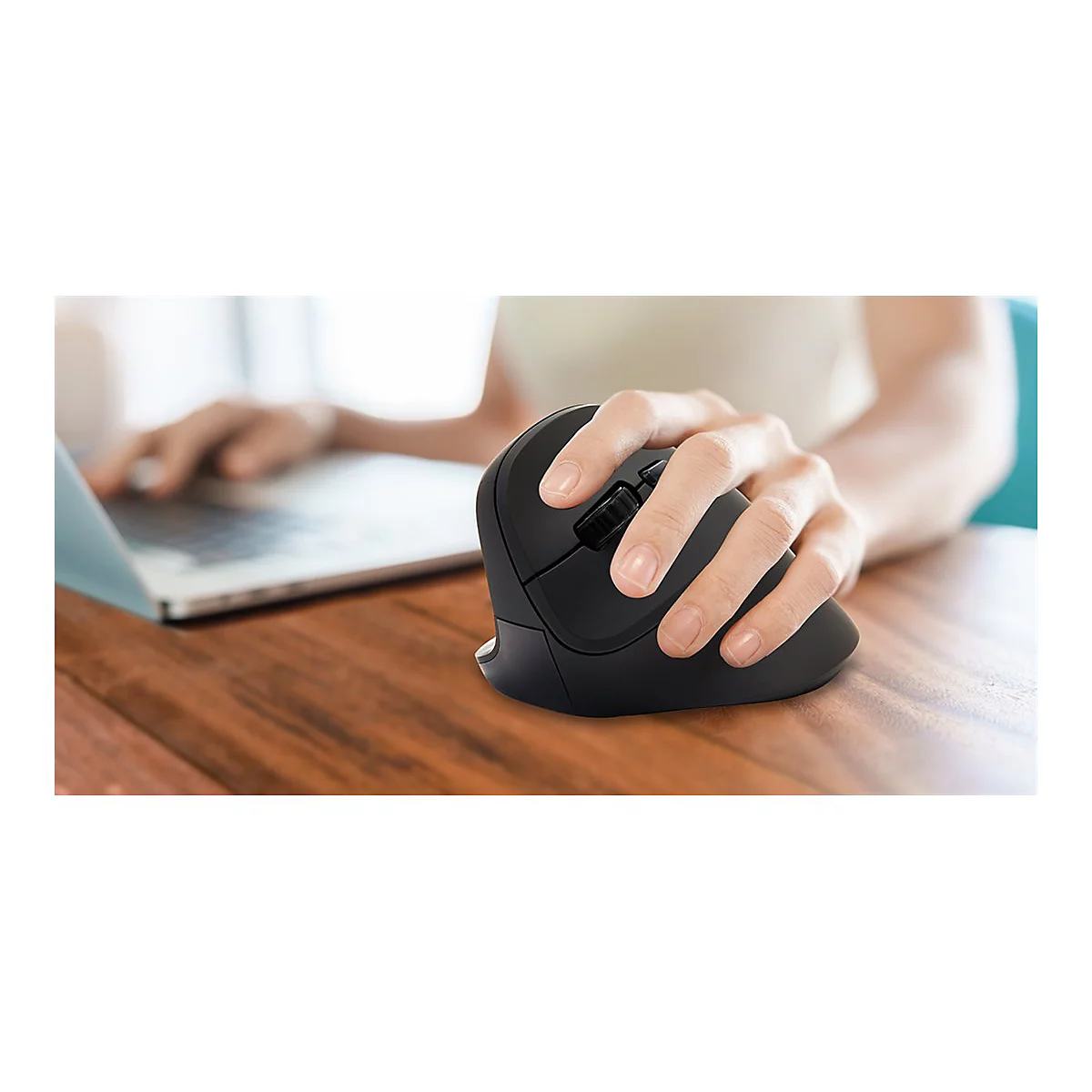Eine Person benutzt eine ergonomische Maus. Die Hand liegt auf der Maus, neben einem Laptop auf einem Holztisch.