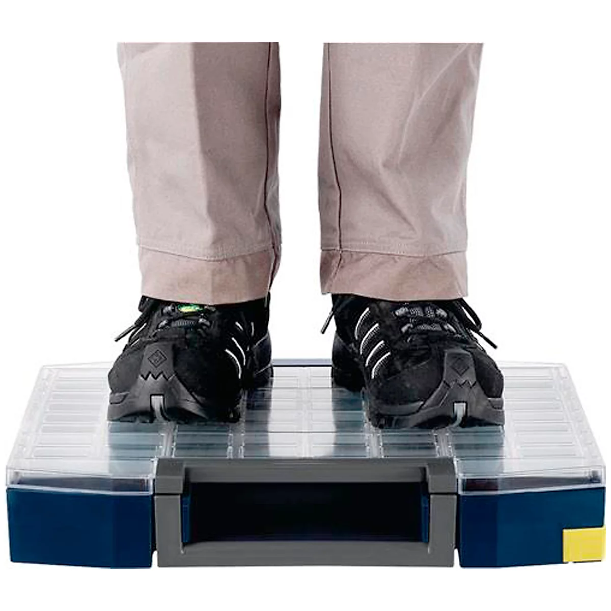 Une personne se tient debout sur une boîte à outils transparente. Chaussures noires, pantalon beige. Boîte à outils avec des accents bleus et jaunes.