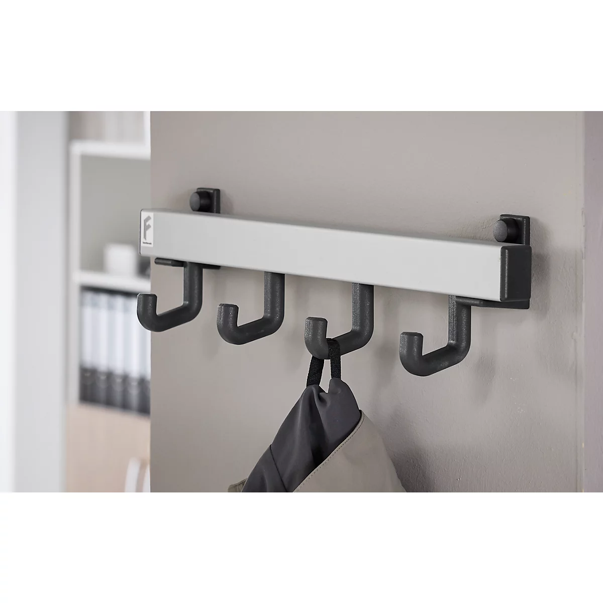 Wandgarderobe met vier haken, waaraan een jas hangt. Daarachter een grijze muur en een plank.