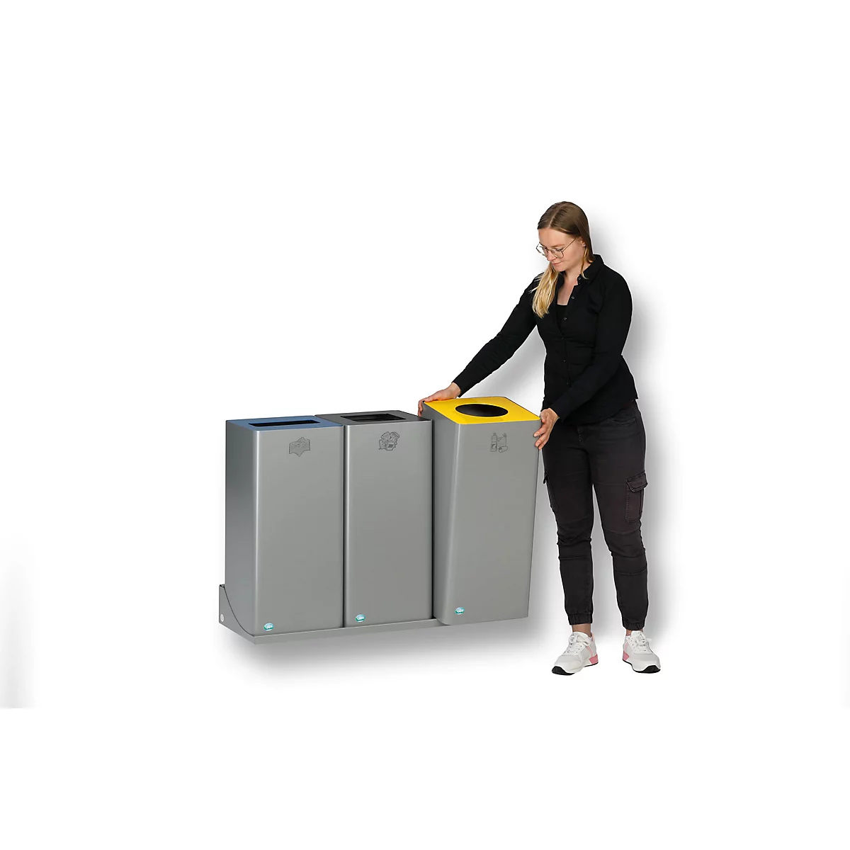 Eine Frau steht neben drei Mülltonnen. Eine Tonne hat einen gelben Deckel, die anderen haben blaue und schwarze Deckel.