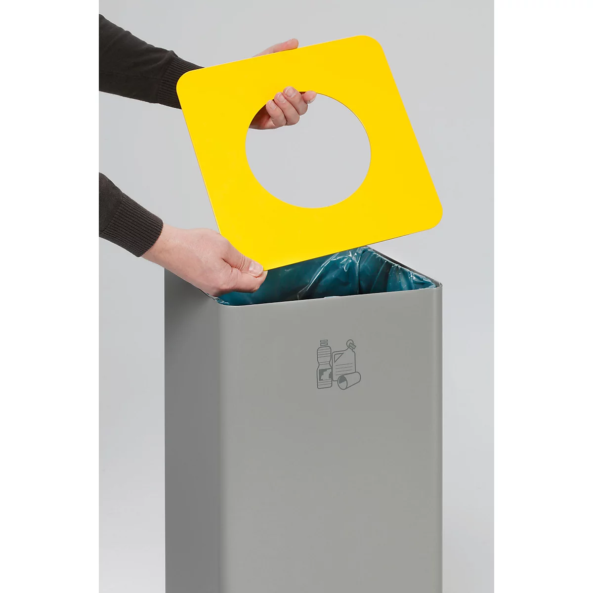 Een persoon houdt een geel vierkant deksel met een rond gat boven een grijze vuilnisbak.
