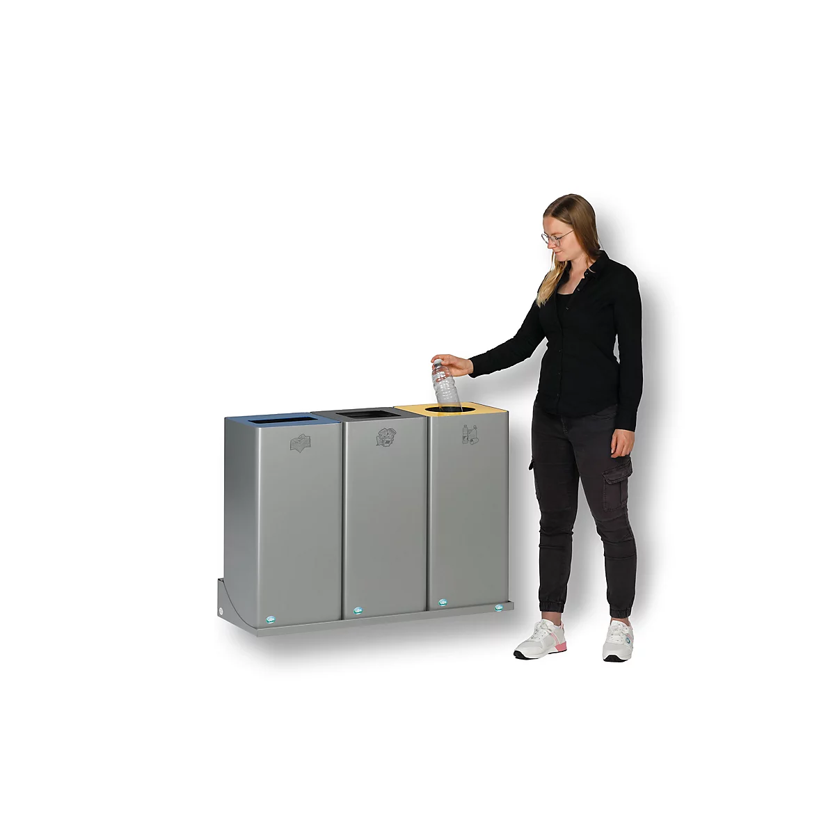 Vrouw die glas in een afvalbak gooit. Drie grijze bakken met gekleurde deksels: blauw, zwart en geel.