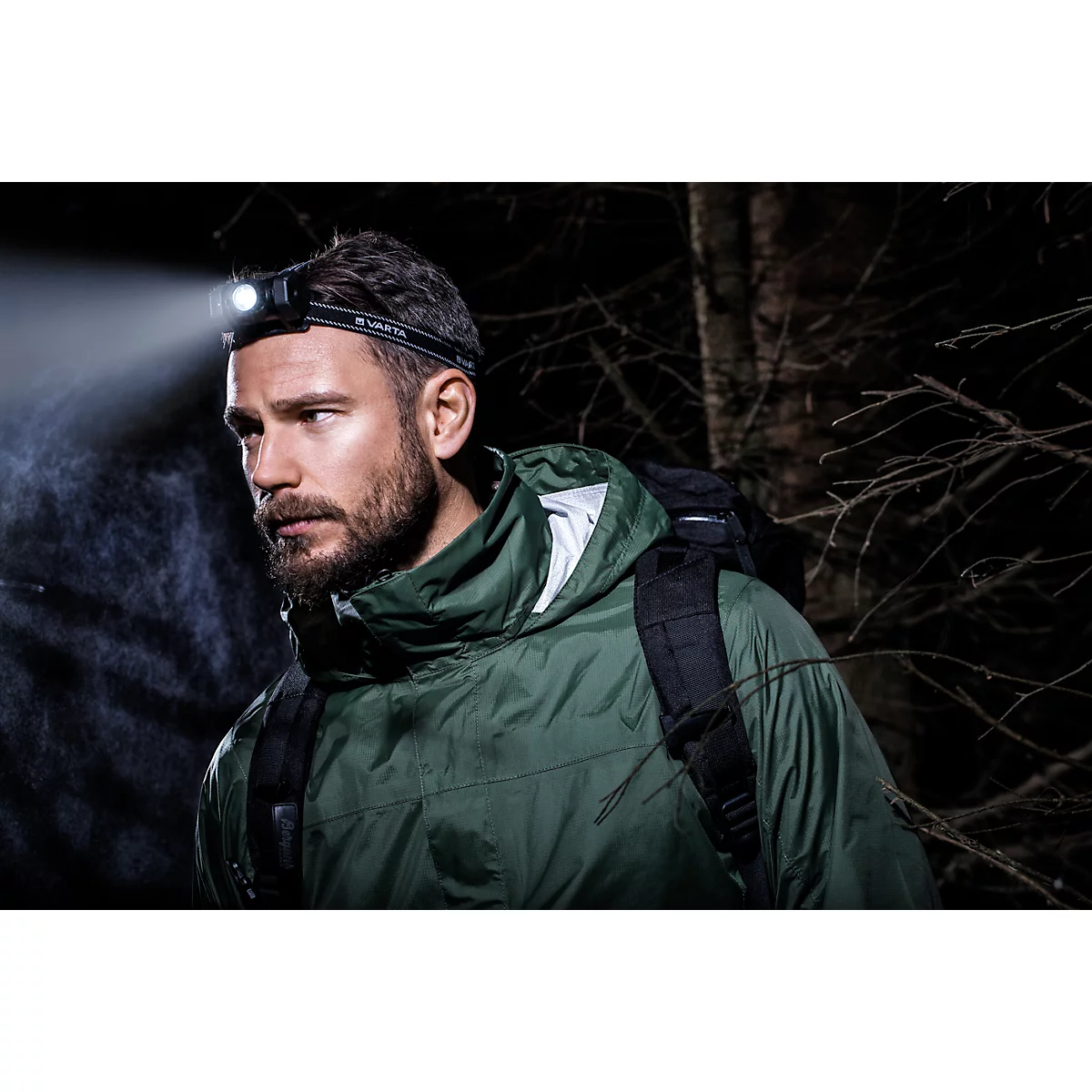 A man in the dark wearing a headlamp. He is wearing a jacket and a backpack.