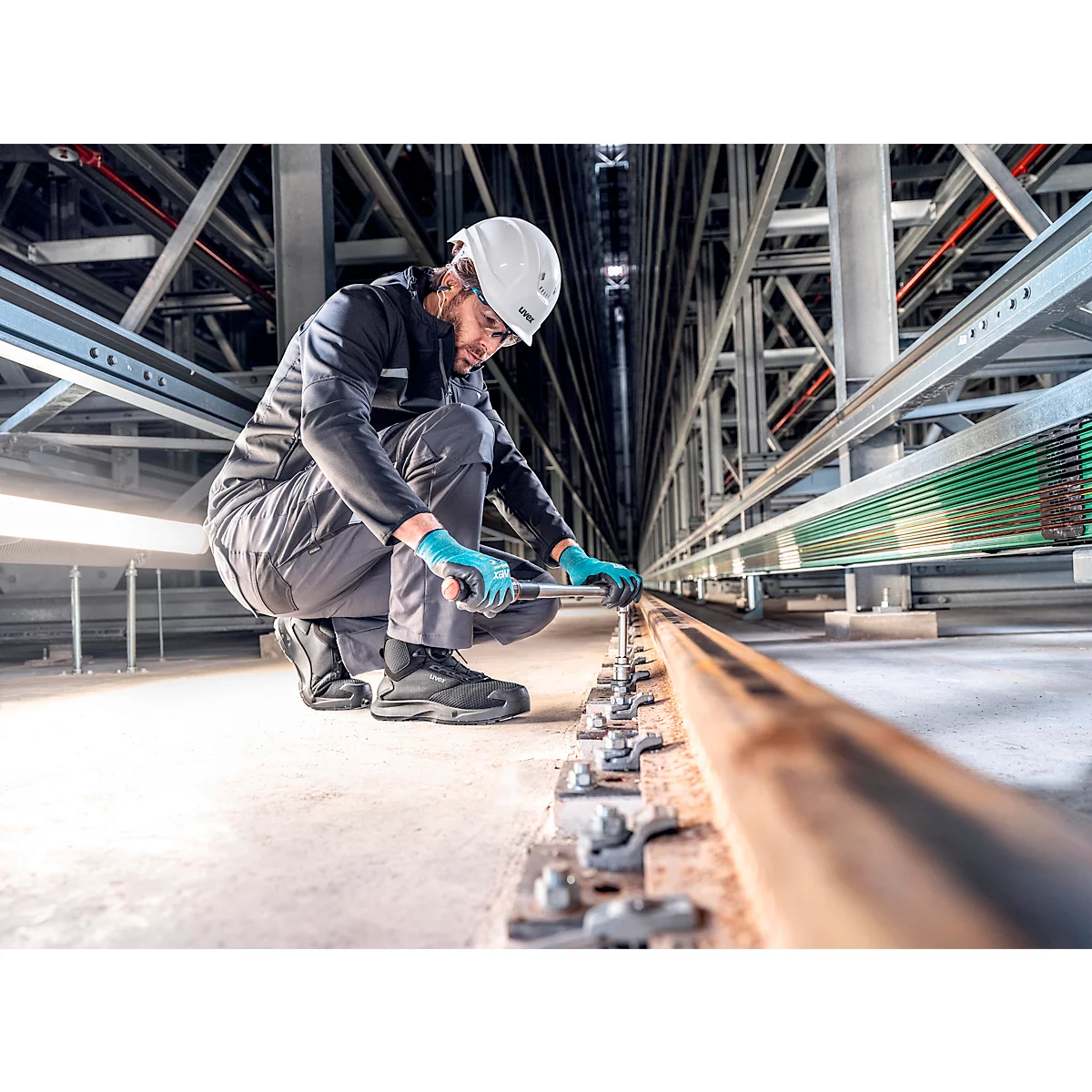 Een man in werkkleding knielt bij een rail, houdt gereedschap vast en schroeft. Hij draagt een helm, handschoenen.