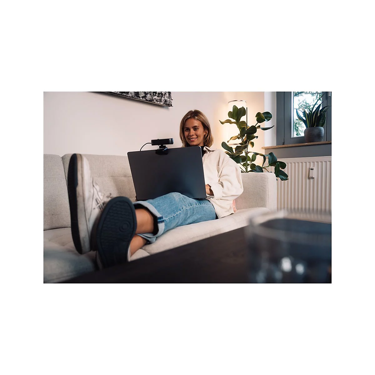 Eine lÃ¤chelnde Frau sitzt entspannt auf einem Sofa mit Laptop und Webcam.