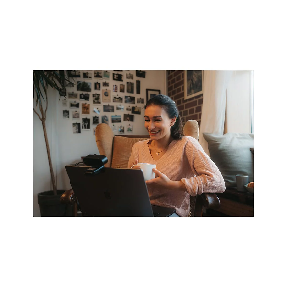Eine lÃ¤chelnde Frau sitzt vor einem Laptop und hÃ¤lt eine Tasse. Im Hintergrund sind Fotos an der Wand und eine Topfpflanze zu sehen.