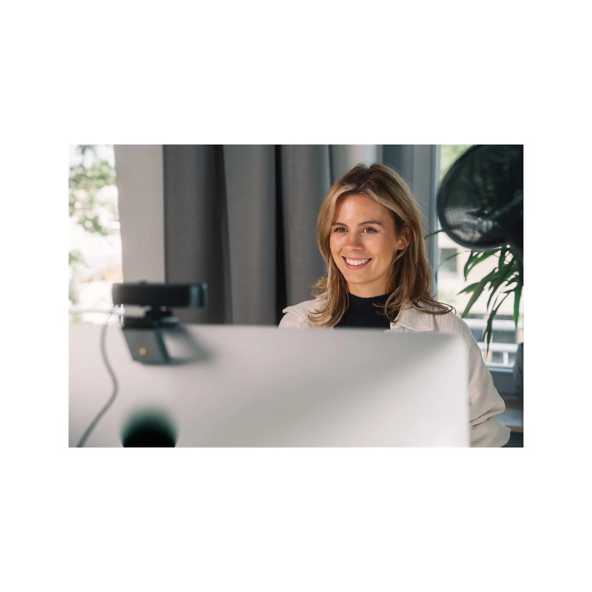 Eine lÃ¤chelnde Frau sitzt vor einem Computer mit Webcam. Helle Kleidung, blondes Haar, Vorhang im Hintergrund.