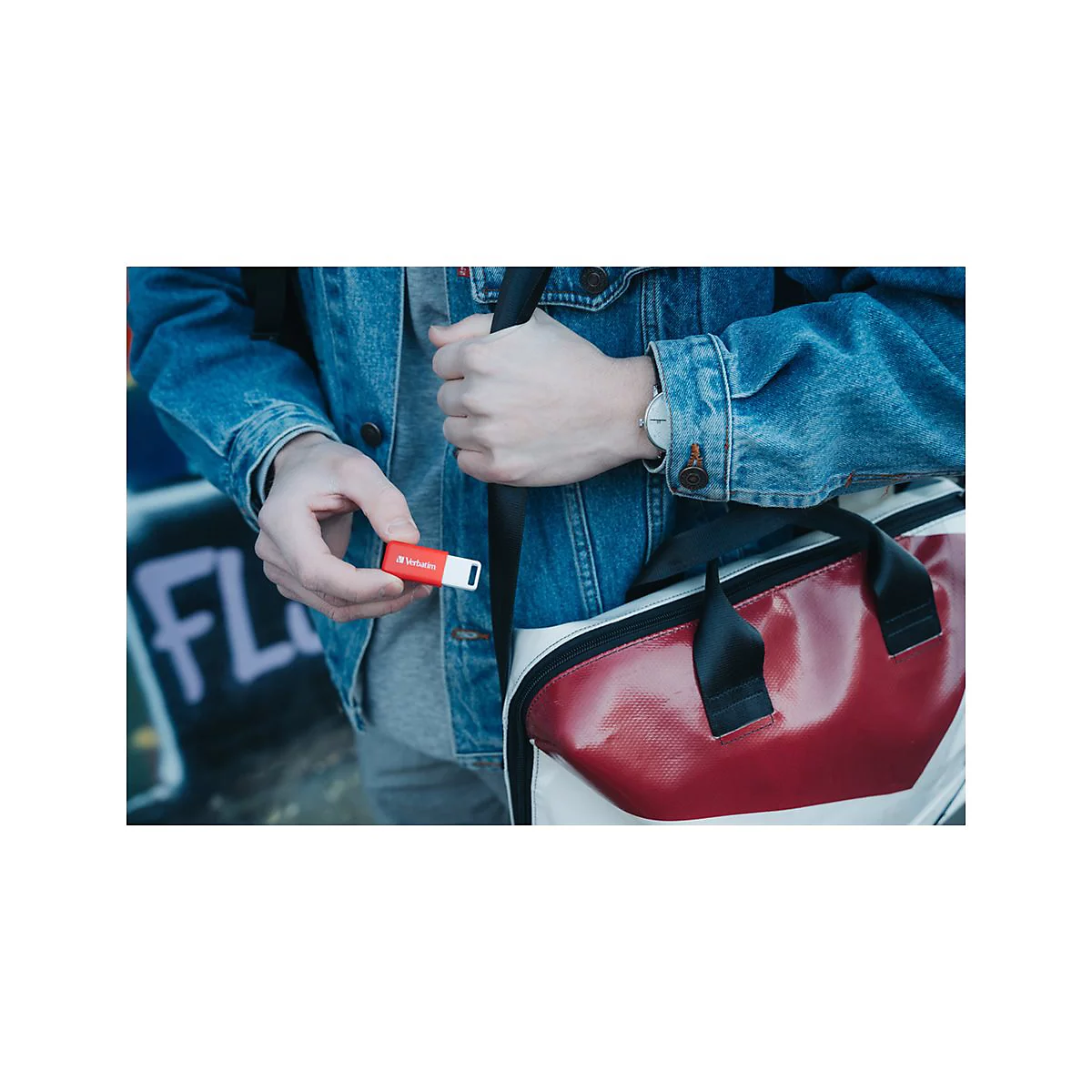Person hÃ¤lt roten USB-Stick. In der anderen Hand eine Tasche. Denim-Jacke und Uhr.