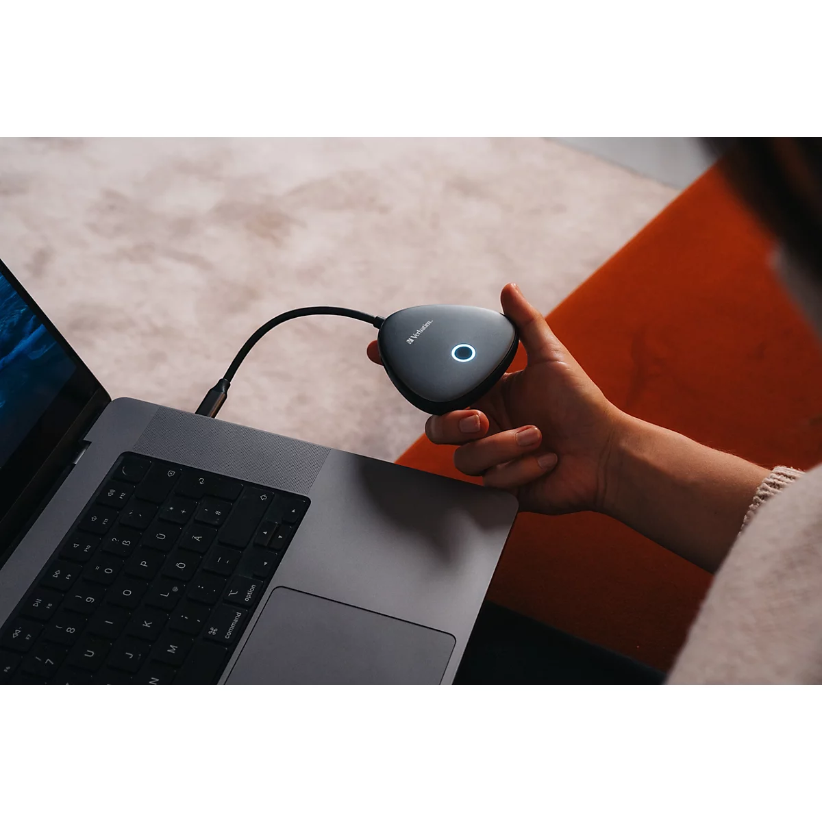 Person hält einen dreieckigen, grauen Barco ClickShare Button mit leuchtendem Ring an einem Laptop.