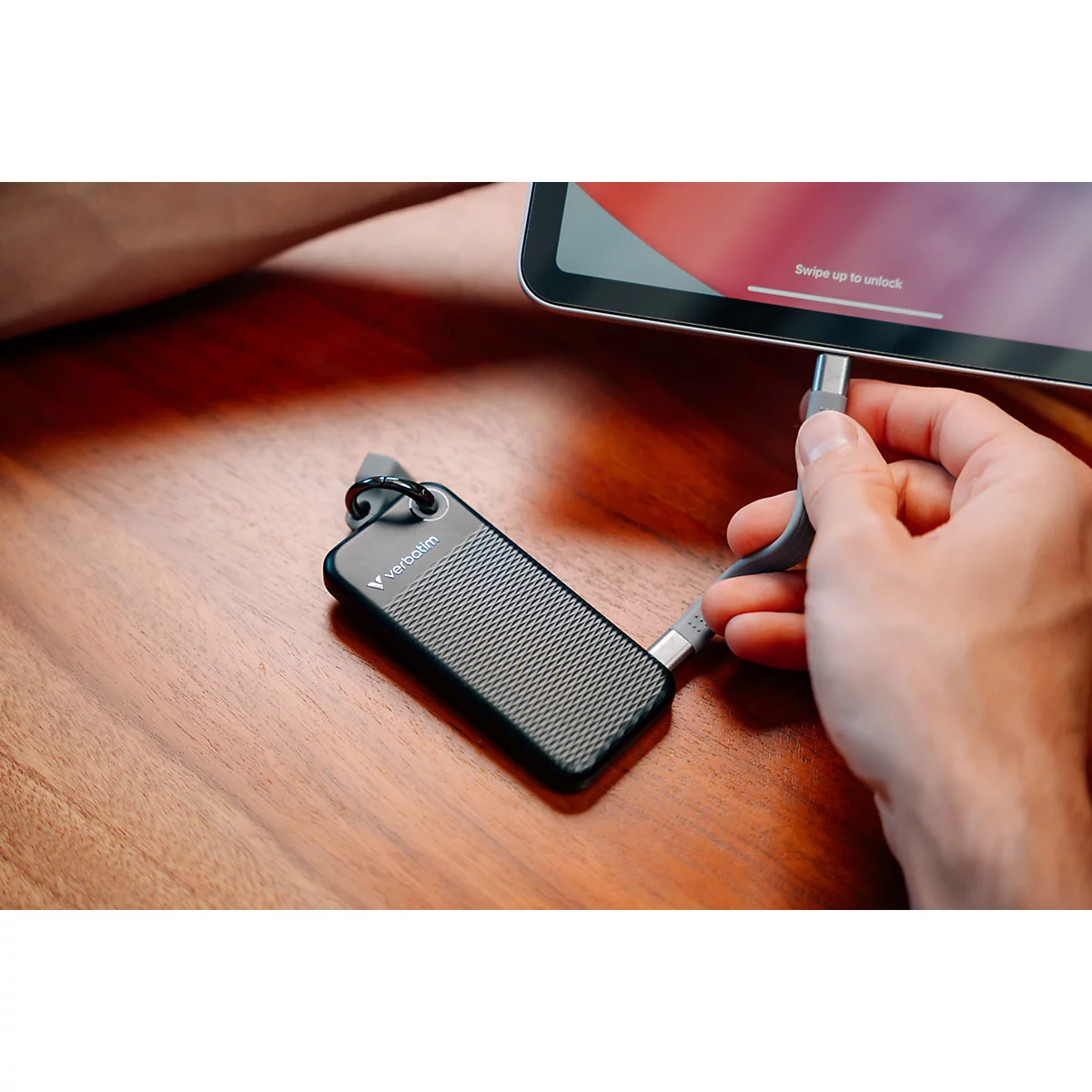 Eine Hand schließt eine kleine schwarze Verbatim-SSD mit kurzem USB-C-Kabel an ein Tablet auf einem Holztisch an.