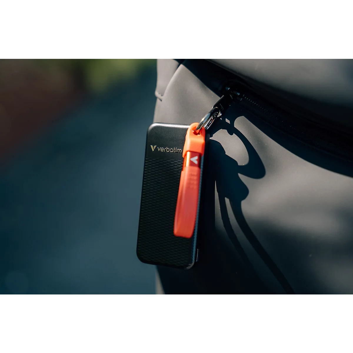 Schwarze Verbatim SSD mit orangefarbenem Karabinerband, befestigt an einer grauen Tasche.