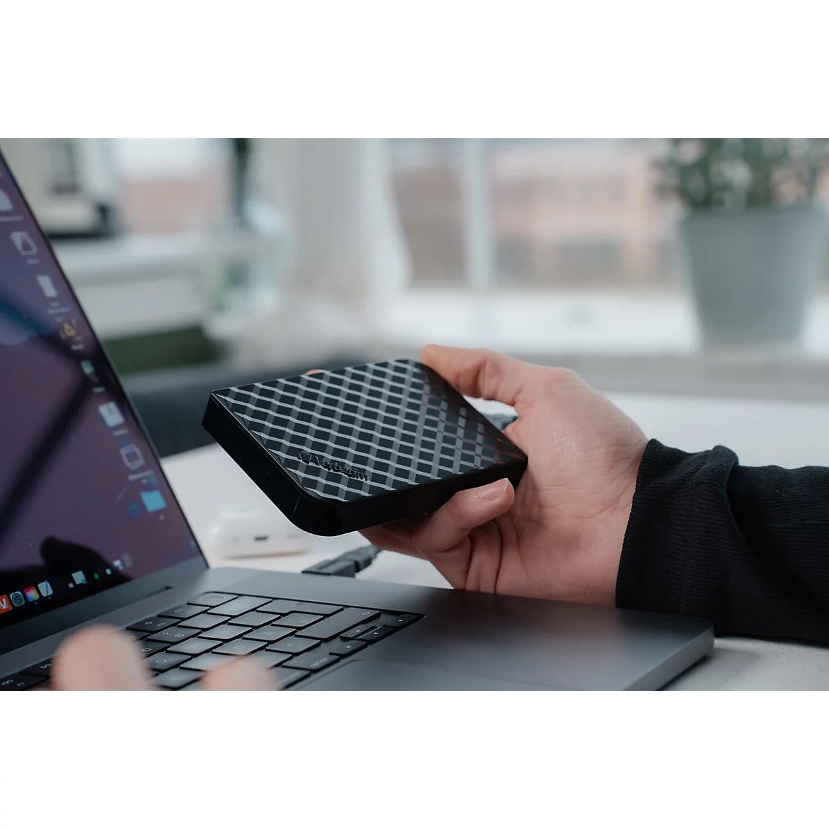 Eine Hand hält eine schwarze, texturierte externe Festplatte neben einem Laptop an einem hellen Schreibtisch.