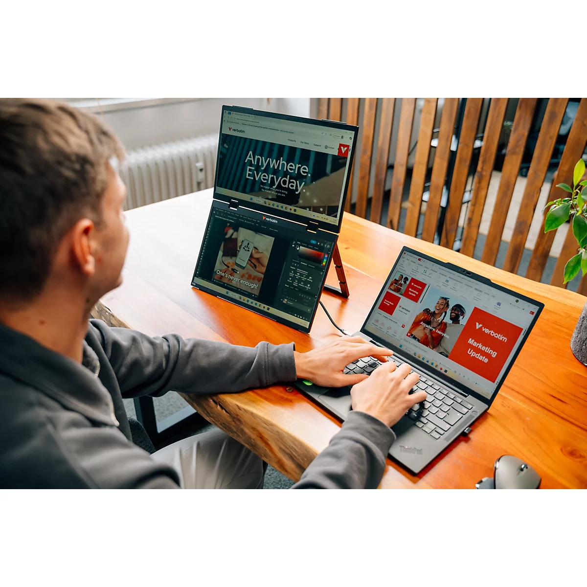 Person arbeitet an einem Laptop mit angeschlossenem zweiten Bildschirm auf einem Holztisch.
