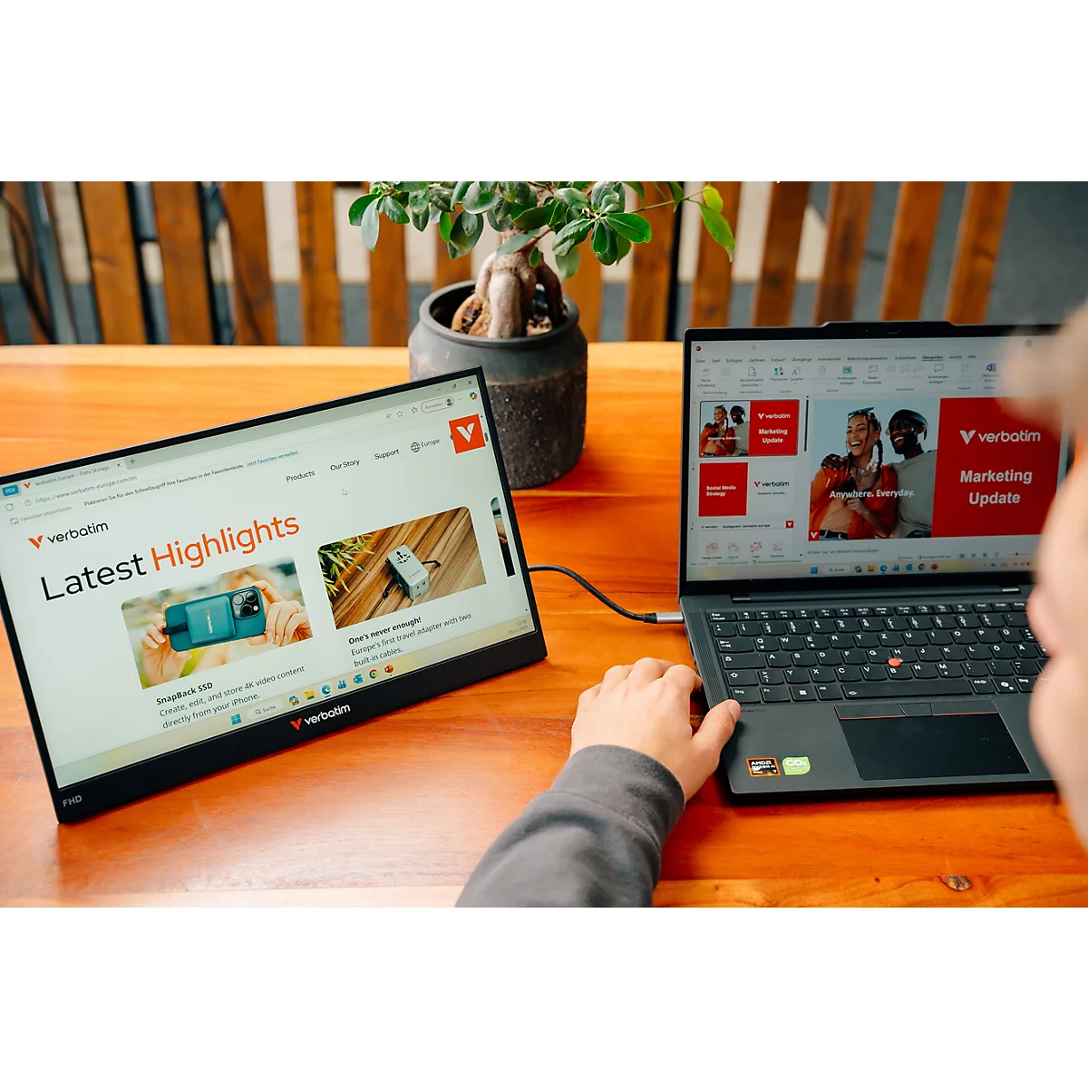 Person arbeitet an einem Laptop mit angeschlossenem externen Verbatim-Monitor auf einem Holztisch.