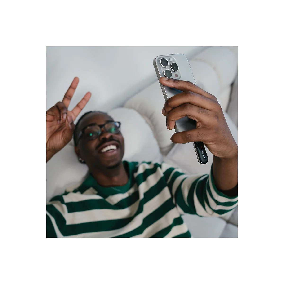 Ein Mann mit Brille macht ein Selfie. Er zeigt das Victory-Zeichen und lächelt. Er hält ein Handy in der Hand.