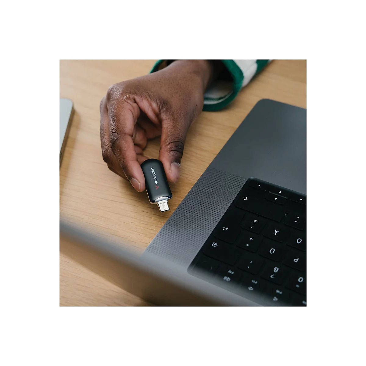 Eine dunkle Hand hält einen schwarzen USB-Stick mit der Aufschrift 'verbatim'. Er ist über einem grauen Laptop platziert.