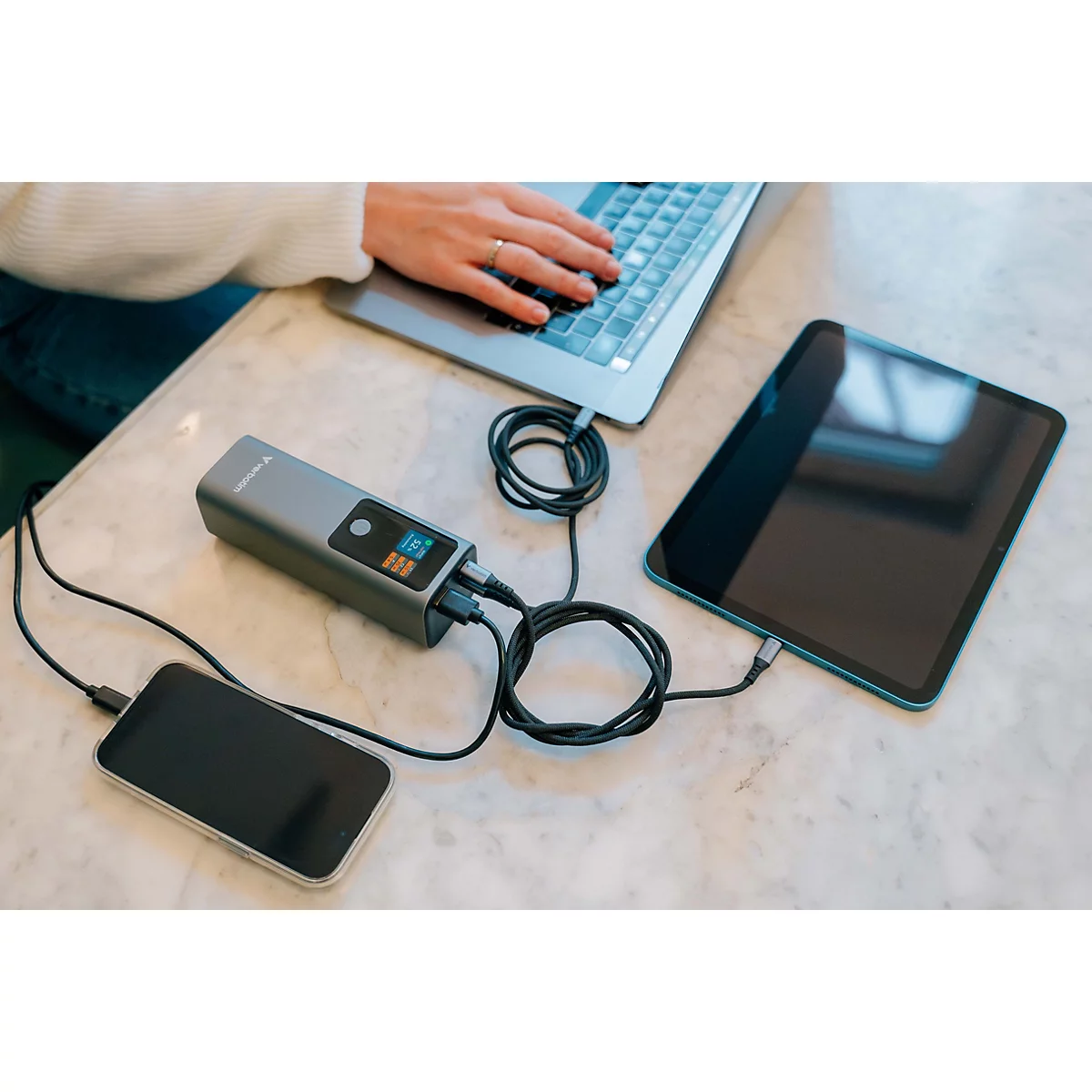 Eine graue Powerbank lädt gleichzeitig einen Laptop, ein Tablet und ein Smartphone auf einem hellen Tisch.