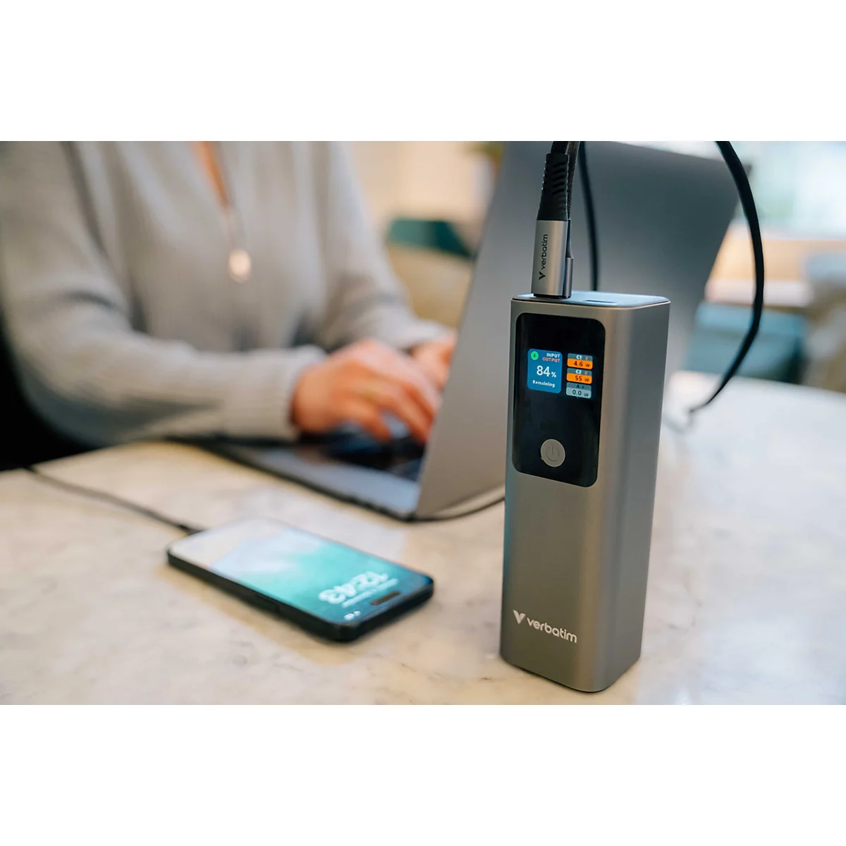 Eine graue Verbatim Powerbank steht auf einem Tisch und lädt ein Smartphone, während im Hintergrund jemand am Laptop arbeitet.
