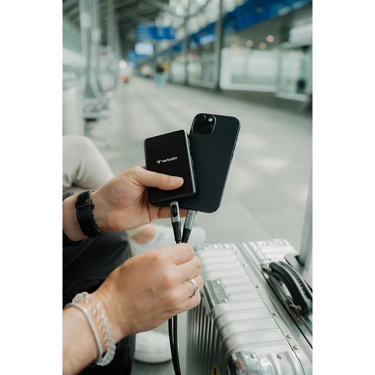 Person lädt ein schwarzes Smartphone mit einer schwarzen Powerbank an einem Bahnhof.