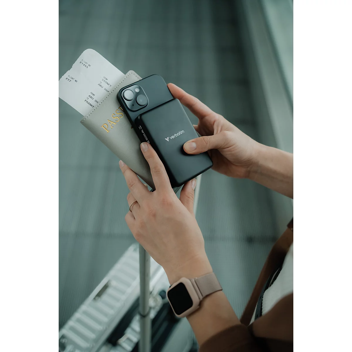 Hände halten eine Powerbank, ein Smartphone und einen Reisepass mit Bordkarte an einem Flughafen.