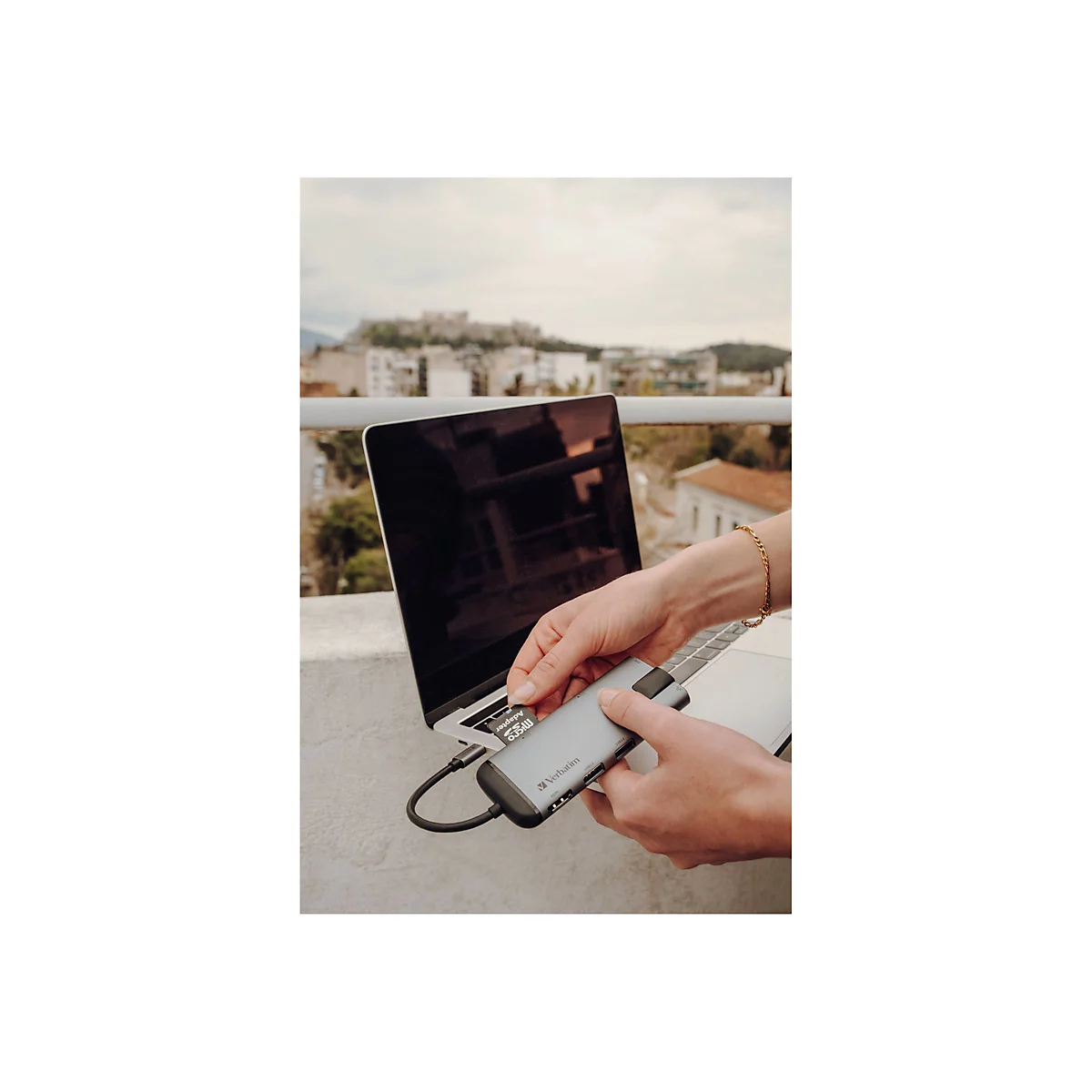 Une personne insère une carte SD dans un adaptateur USB-C tout en travaillant sur un ordinateur portable. Des bâtiments et une ville sont visibles en arrière-plan.