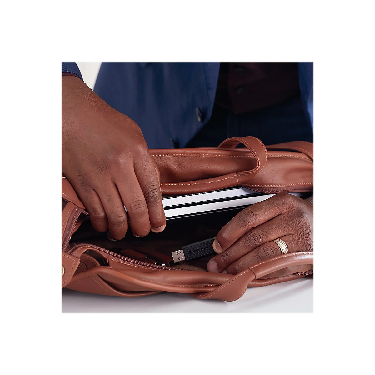 Mains plongeant dans un sac en cuir marron. Ã€ l'intÃ©rieur, un ordinateur portable, une clÃ© USB et d'autres objets.