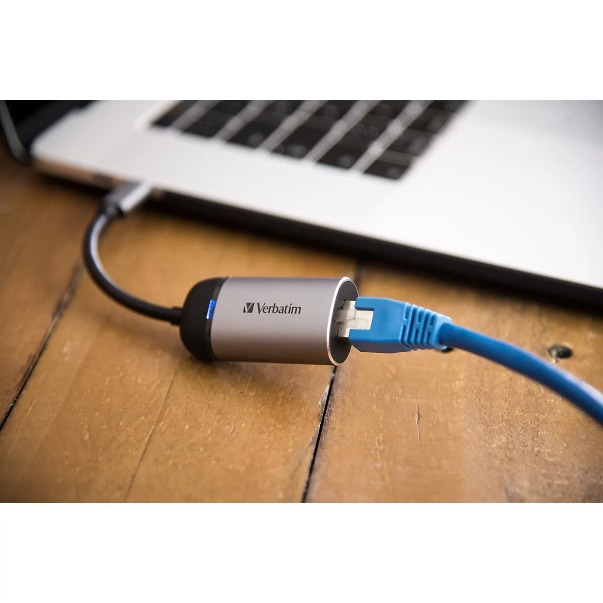 Ein silberner Verbatim-Ethernet-Adapter, verbunden mit einem Laptop und einem blauen Netzwerkkabel auf einem Holztisch.