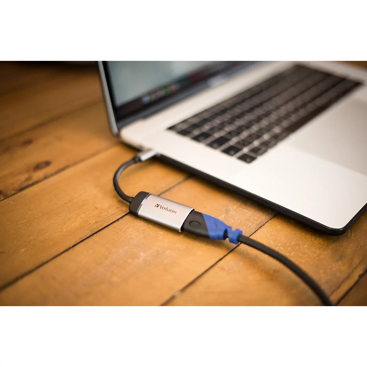 Ein silberner Verbatim-Adapter ist an einem Laptop angeschlossen und liegt auf einem hölzernen Tisch.