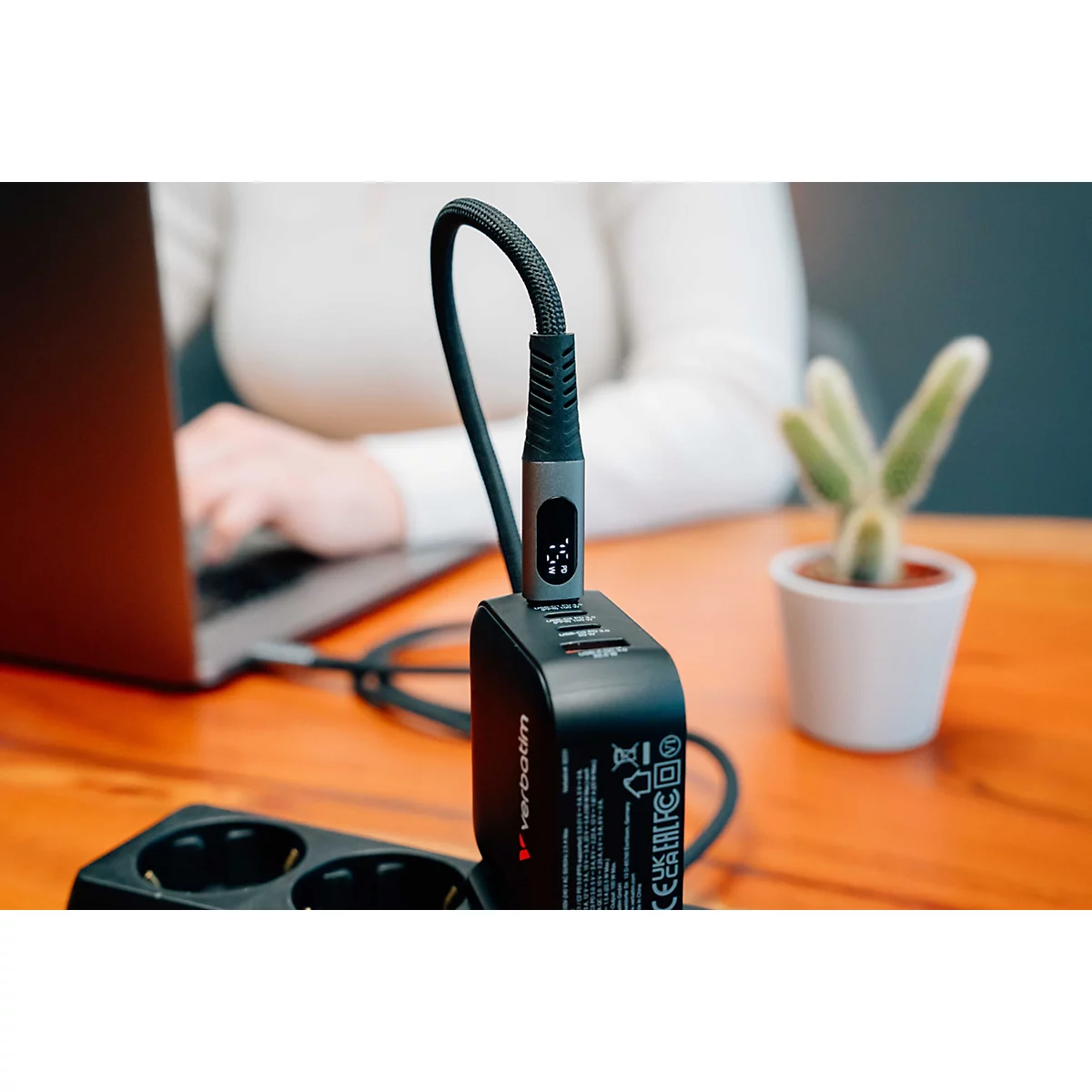 Schwarzes Verbatim-Ladegerät mit USB-C-Kabel in einer Steckdose auf einem Schreibtisch bei der Arbeit.