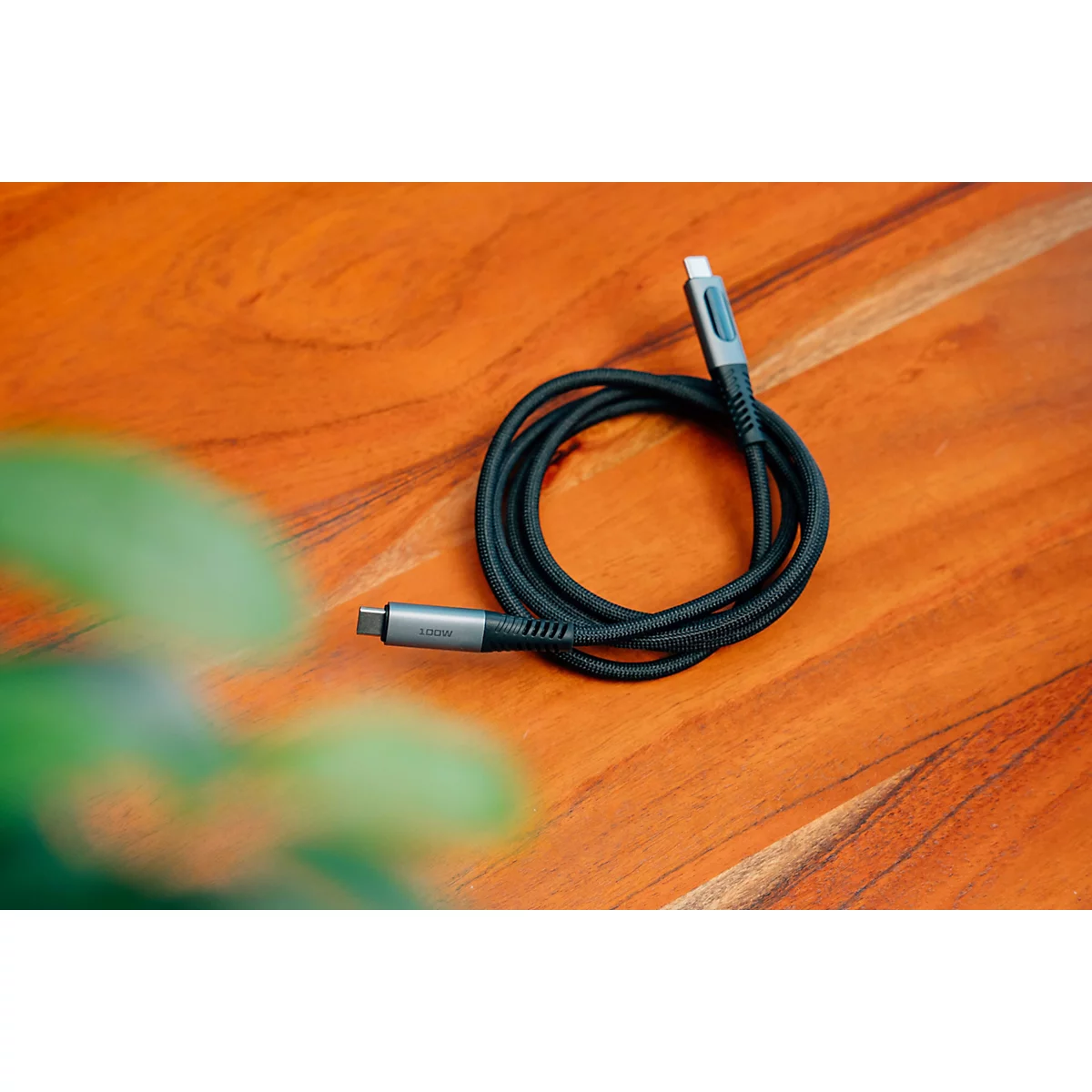 Ein aufgerolltes schwarzes USB-C-Kabel liegt auf einem hölzernen Tisch, unscharfe grüne Pflanzen im Vordergrund.